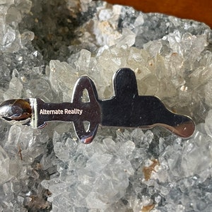 May include: A silver-colored metal pin with the words "Alternate Reality" engraved on it. The pin is shaped like a stylized letter "A" with a circle in the middle.