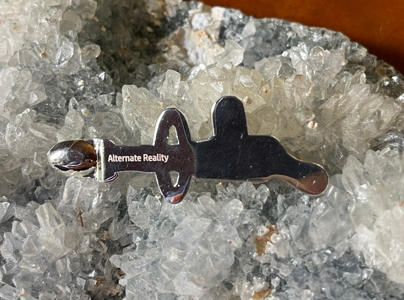 May include: A silver-colored metal pin with the words "Alternate Reality" engraved on it. The pin is shaped like a stylized letter "A" with a circle in the middle.