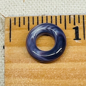 May include: A dark blue, donut-shaped bead with white and purple swirls. The bead is approximately 1 inch in diameter and sits on a wooden ruler. The bead has a glossy finish.