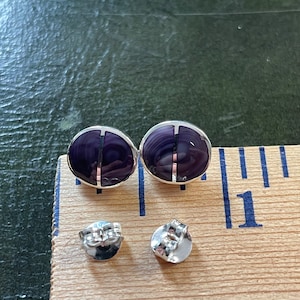 May include: A pair of silver stud earrings with purple abalone shell inlays. The earrings are shown on a wooden surface with a ruler.