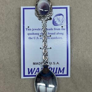 May include: A decorative spoon with a silver-colored handle and a purple and white oval-shaped stone at the top. The spoon is attached to a white card with text that reads "This jewelry made from the quahaug shell found along the U.S. eastern seashore."