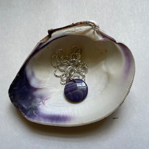 May include: A silver chain necklace with a purple and white swirl pendant resting inside a white seashell.