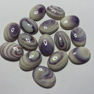 May include: A collection of oval-shaped cabochons in shades of lavender, purple, and cream. The stones have a polished, glossy finish and feature swirling patterns. The cabochons are arranged on a white surface.