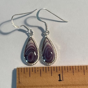 May include: A pair of silver drop earrings featuring teardrop-shaped stones with concentric purple and white bands. The earrings are positioned above a ruler showing a scale in inches, with the stones measuring approximately 0.5 inches in length.