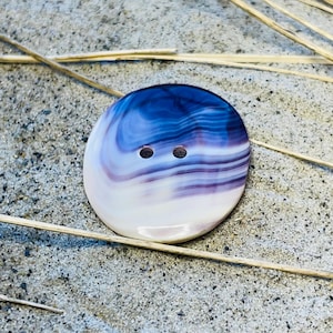 May include: A round, white and purple shell button with two holes. The button has a swirling pattern.