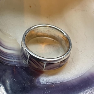 May include: A silver ring with a band of rectangular purple and white stones. The ring is set against a backdrop of a purple and white seashell, creating a contrast of textures and colors. The ring's design is simple yet elegant.