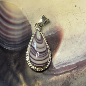 May include: A silver pendant with a teardrop-shaped purple and white shell inlay. The pendant has a rope-like border around the shell.