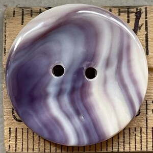 May include: A large, round button with a marbled design in shades of purple and white. The button has two holes and a glossy finish. The button is placed on a wooden ruler.