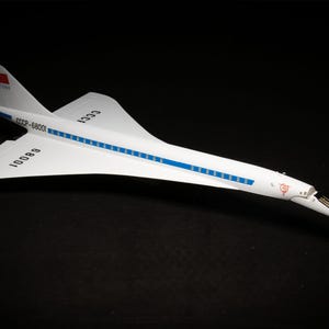 Pode incluir: Um modelo branco do avião de passageiros supersônico soviético Tupolev Tu-144, também conhecido como 'Concordski'. O modelo tem o número de registro 'CCCP-68001' e o texto 'CCCT' na fuselagem. O modelo está sobre um fundo preto.