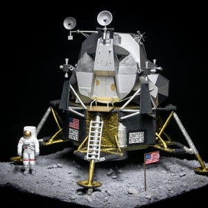 Op de afbeelding: Een model van de Apollo Lunar Module, ook bekend als de Lunar Excursion Module of LEM, met een klein astronautfiguur er voor. Het model staat op een grijs oppervlak dat lijkt op het oppervlak van de maan. De LEM is zilver en zwart met een gouden basis en een ladder aan de zijkant. De Amerikaanse vlag is in het maanoppervlak geplant.