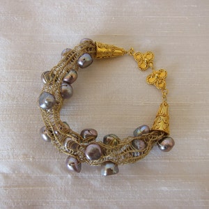 May include: A gold and gray pearl bracelet with a crocheted design. The bracelet features gold accents and a clasp.