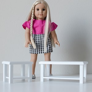 May include: Two white wooden doll-sized coffee tables. The table on the right has a shelf. A doll wearing a pink and black checkered dress stands between the tables.