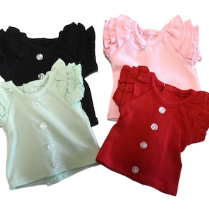 May include: Four short-sleeved shirts in black, light green, light pink, and red. Each shirt features a round neckline, layered ruffled sleeves, and a vertical row of four clear, circular buttons down the center front of the garment.