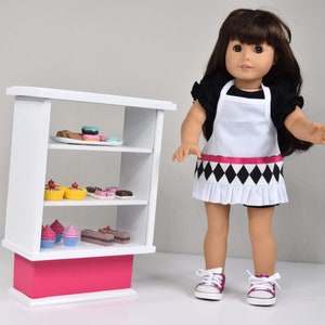 May include: A doll with long brown hair wearing a black shirt, a white apron with a black and white diamond pattern and a pink ribbon, and pink sneakers. Beside the doll is a white three-tiered display shelf with a pink base holding miniature food.