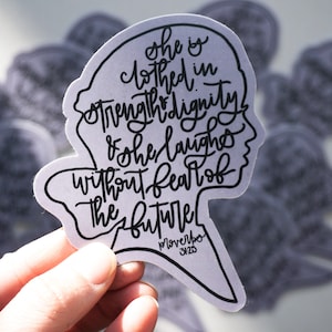 Könnte beinhalten: Ein weißer Aufkleber mit einer schwarzen Silhouette eines Frauenkopfes und dem Text "Sie ist bekleidet mit Stärke und Würde und sie lacht ohne Angst vor der Zukunft" und dem Datum "19. November 3425".