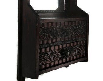 May include: A dark brown wooden wall-mounted mirror featuring a rectangular glass pane framed by beaded detailing. Below the mirror is a storage compartment with a front panel decorated with intricate carved floral patterns and horizontal lines.