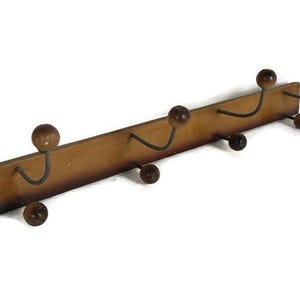 Può includere: Un appendiabiti da parete in legno con cinque ganci in metallo. I ganci hanno pomelli in legno alle estremità. L'appendiabiti è tinto di un colore marrone scuro.