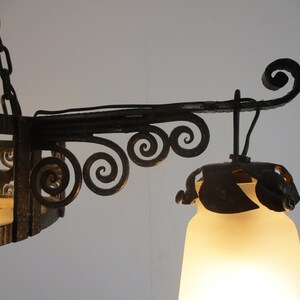 May include: Close-up of a wrought iron chandelier with a frosted glass shade. The fixture features decorative scrollwork and a chain for hanging. The light is on, illuminating the frosted glass shade.