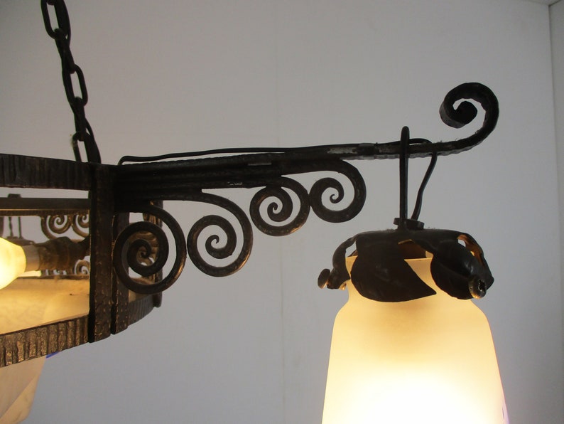 May include: Close-up of a wrought iron chandelier with a frosted glass shade. The fixture features decorative scrollwork and a chain for hanging. The light is on, illuminating the frosted glass shade.