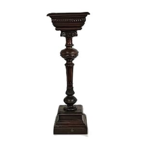 May include: A dark brown, ornate wooden plant stand with a square base and a decorative top. The stand features a detailed, carved design and a tall, slender column. The top is a small, square tray, perfect for displaying a small plant or decorative item.