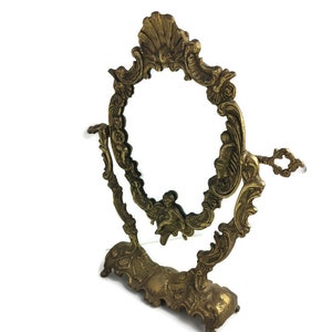 Puede incluir: Un espejo de tocador de mesa dorado y adornado con un soporte giratorio. El espejo tiene un marco decorativo de forma ovalada con detalles intrincados.
