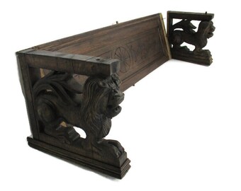 May include: Dark brown wooden shelf with carved lion supports. The shelf has a decorative carved design on the front. The lion supports are intricately carved with wings and detailed features. The shelf is angled, showing its construction.