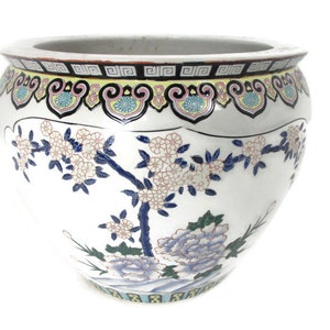 May include: A white ceramic planter with a blue and pink floral design. The planter has a wide mouth and a rounded body. The design features a tree with flowers and leaves, as well as a large flower at the bottom.