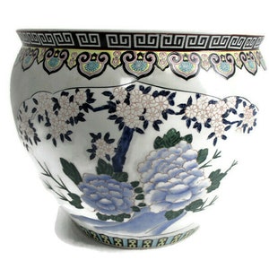 May include: A large white ceramic planter with a blue and white floral design. The planter has a wide mouth and a rounded body. The floral design features blue and white flowers, green leaves, and a blue and white tree.