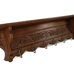 May include: A dark brown wooden wall shelf with ornate carved floral detailing. The shelf has a series of six brass-colored hooks along the bottom edge. The shelf is designed for storage and hanging items.