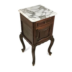 May include: A dark wood bedside table with a white marble top and a single drawer. The bedside table has ornate legs and a carved door.