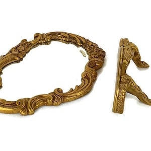 May include: A gold-toned ornate oval mirror with a decorative stand. The mirror has a detailed, intricate design with floral motifs. The stand is also gold-toned and has a similar ornate design.
