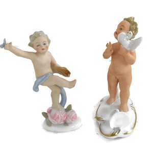 Puede incluir: Dos figuritas de porcelana de querubines. Un querubín sostiene una paloma blanca y está de pie sobre una nube blanca con ribetes dorados. El otro querubín sostiene una mariposa azul y está de pie sobre una base con rosas rosas.