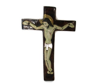 May include: A wooden crucifix with a dark brown cross and a painted figure of Jesus. The figure has a green-toned body, a white loincloth, and a gold halo. The cross is mounted on a white background.