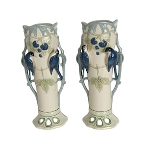 Op de afbeelding: Set van twee crèmekleurige keramische vazen met blauwe vogel- en bessenontwerpen. De vazen hebben lichtblauwe randen en accenten, met een decoratieve opengewerkte basis. De vogels zijn diepblauw. Ideaal voor interieurdecoratie.