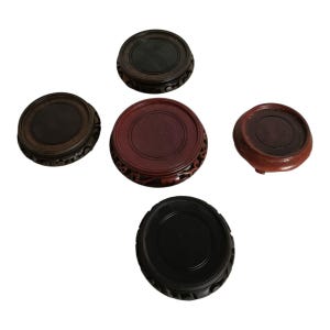 May include: Five wooden display stands in various shades of brown and black. Each stand is round with a recessed center and decorative carved edges. The stands vary in size and wood tone, suitable for displaying small objects.