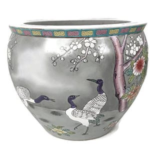 May include: A gray ceramic planter with a wide rim, featuring a hand-painted scene of cranes, flowers, and a tree. The rim has a decorative border with green, pink, and yellow accents. The planter has a rounded shape.