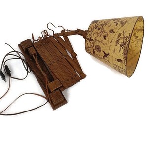 Puede incluir: Una lámpara de pared de madera con una pantalla de tela marrón con un diseño de mapa del mundo vintage. La lámpara tiene un brazo plegable y un cable con un enchufe.