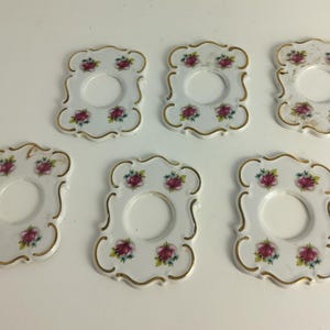 Può includere: Sei sottobicchieri in porcellana bianca con bordo smerlato e finiture dorate. Ogni sottobicchiere presenta un motivo floreale con fiori rosa e verdi. I sottobicchieri hanno un'apertura circolare al centro.