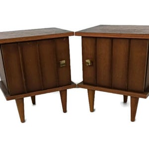May include: A pair of mid-century modern nightstands with wood grain finish and brass hardware. Each nightstand has a single door and four legs.