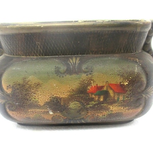 Op de afbeelding: Kleine, antieke keramische plantenbak met een landschapsscène. De plantenbak is bruin met een getextureerde rand en handvatten. Het landschap toont een huis met een rood dak, bomen en een brug. Decoratieve krullen accentueren de scène.