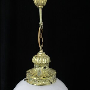 May include: Close-up of a vintage ceiling light fixture. The fixture features a white, rounded glass shade and ornate brass-colored metal components. The metal parts include a decorative ceiling mount, a chain, and a base that holds the glass shade.