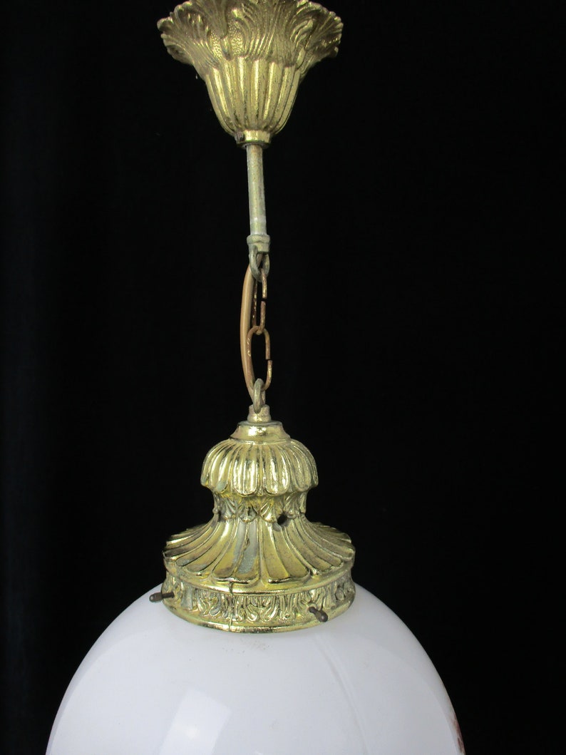 May include: Close-up of a vintage ceiling light fixture. The fixture features a white, rounded glass shade and ornate brass-colored metal components. The metal parts include a decorative ceiling mount, a chain, and a base that holds the glass shade.