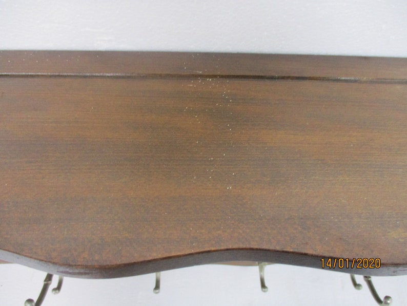 May include: A dark brown wooden shelf with a curved lower edge and several metal hooks. The shelf has a smooth surface and is mounted against a white wall. The shelf is designed for hanging items.