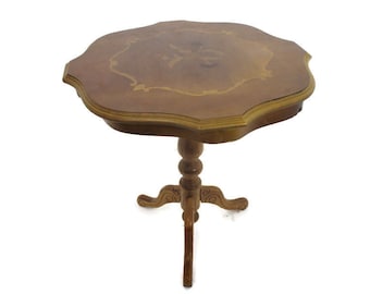 Op de afbeelding: Een vintage, bruine bijzettafel met drie poten en een unieke, geschulpte rand. Het tafelblad heeft een decoratief inlegontwerp. De centrale steun van de tafel heeft een gedraaid, bolvormig ontwerp. De tafel is ongeveer 71 cm hoog.