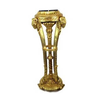 Puede incluir: Un soporte para plantas dorado con una parte superior de mármol negro. El soporte tiene una columna torcida y detalles ornamentados. Es una pieza decorativa para un hogar o jardín.