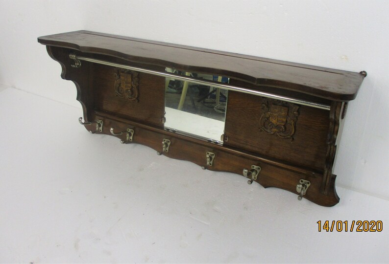May include: A dark brown wooden wall shelf with a mirror, coat hooks, and a metal bar. The shelf has decorative carvings and a classic design, suitable for a hallway or entryway. The shelf measures approximately 100 cm in length.