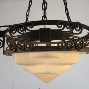May include: An antique chandelier with a tiered, frosted glass shade and ornate, wrought iron detailing. The fixture is suspended by chains and features decorative scrollwork and a dark finish. A classic lighting fixture.