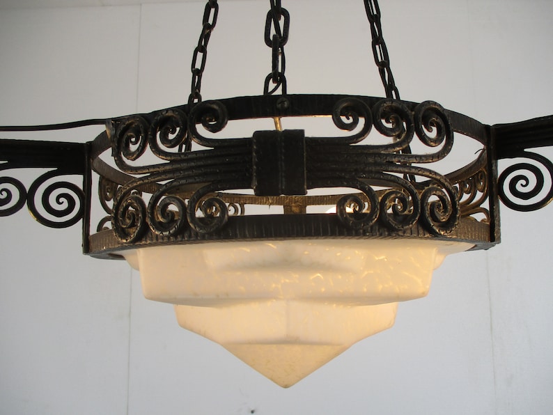 May include: An antique chandelier with a tiered, frosted glass shade and ornate, wrought iron detailing. The fixture is suspended by chains and features decorative scrollwork and a dark finish. A classic lighting fixture.