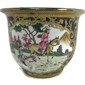 Peut inclure: Un pot de fleurs décoratif en céramique avec une scène de chasse détaillée. Le pot a un bord doré et une bordure noire et dorée. Le motif central représente des personnages à cheval, des chiens, des arbres et une église, le tout dans des couleurs vives.