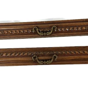 Puede incluir: Dos frentes de cajón rectangulares de madera con tallas ornamentadas y tiradores de latón. La madera tiene un tono marrón cálido. Los frentes de los cajones son probablemente para una cómoda o un armario.